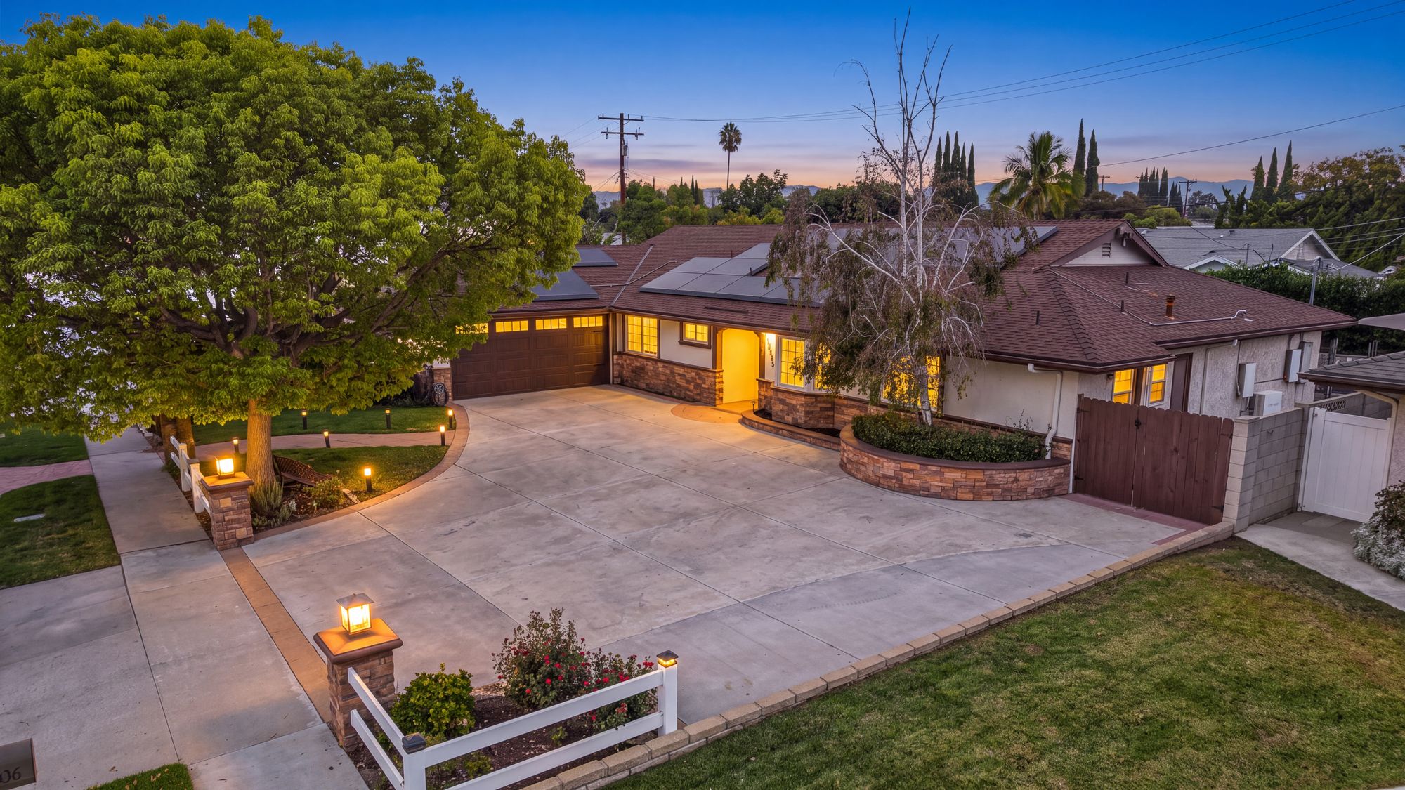 The front of Serenity at dusk: a single-story home with a wide driveway, a shade tree and a white fence